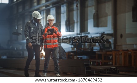 Two Heavy Industry Engineers Walk in Steel Factory, Use Tablet and Discuss Work. Industrial Worker Uses Angle Grinder in the Background. Black African American Specialist Talks to Female Technician. Royalty-Free Stock Photo #1870469452