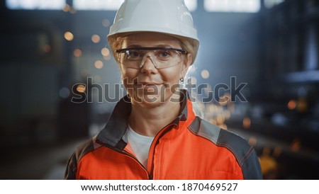 Portrait of a Professional Heavy Industry Engineer Worker Wearing Uniform, Glasses and Hard Hat in a Steel Factory. Beautiful Female Industrial Specialist Standing in Metal Construction Facility. Royalty-Free Stock Photo #1870469527