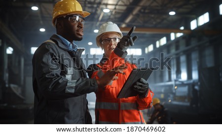 Two Heavy Industry Engineers Stand in Steel Metal Manufacturing Factory, Use Digital Tablet Computer and Have a Discussion. Black African American Industrial Specialist Talk to Female Technician. Royalty-Free Stock Photo #1870469602