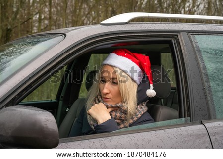 Young blonde woman in a Santa Claus hat is sitting at the wheel of a car and looks with a thoughtful, sad look into the distance