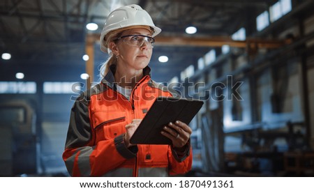 Professional Heavy Industry Engineer Worker Wearing Safety Uniform and Hard Hat Uses Tablet Computer. Serious Successful Female Industrial Specialist Walking in a Metal Manufacture Warehouse. Royalty-Free Stock Photo #1870491361