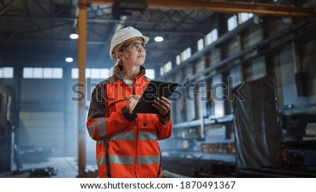 Professional Heavy Industry Engineer Worker Wearing Safety Uniform and Hard Hat, Using Tablet Computer. Serious Successful Female Industrial Specialist Walking in a Metal Manufacture Warehouse. Royalty-Free Stock Photo #1870491367