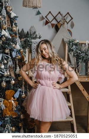 Beautiful blonde with curls stands near a Christmas tree in a pink dress