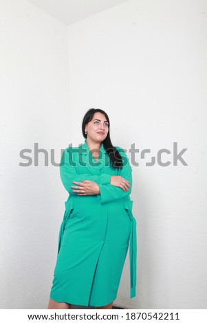 Overweight brunette woman in  summer spring turquoise dress. She is posing standing isolated on white walls background in simple interior. Fashion and style concept. Full length, copy space. Advert