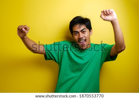 Asian man in green t-shirt smiling and dancing happily, joyful expressing celebrating good news victory winning success gesture against yellow background