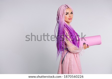 Young girl in pink dress, hair braids and yoga mat looking at the camera against a white background, empty side space.