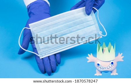Coronavirus image and hands holding medical mask on blue background with copyspace. Hands in gloves hold cloth mask for virus covid-19 protection. Fun cartoon creature.