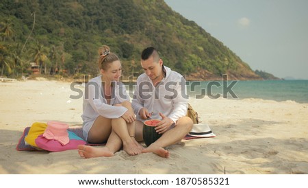 The cheerful love couple holding and eating slices of watermelon on tropical sand beach sea. Romantic lovers two people caucasian spend summer weekend in outdoor. Hat, backpack white shirt beachwear. Royalty-Free Stock Photo #1870585321