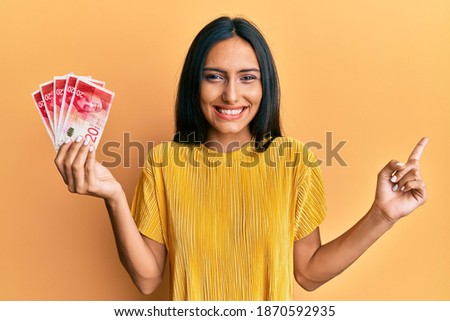 Young brunette woman holding 20 israel shekels banknotes smiling happy pointing with hand and finger to the side 