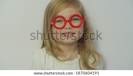 Close-up little blond girl in looking at camera in Red toy glasses stretches smile on white background. childhood, celebration, birthday, games concept.