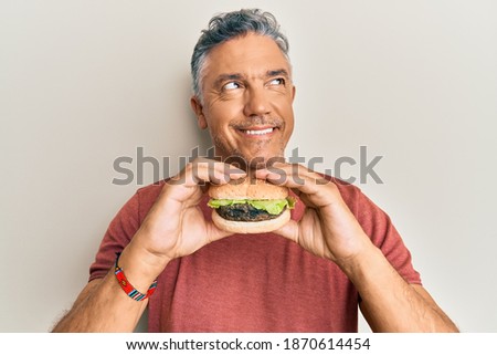Handsome middle age mature man eating a tasty classic burger smiling looking to the side and staring away thinking. 