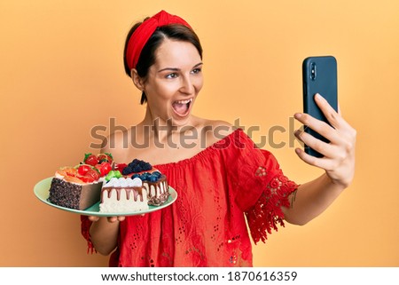 Young brunette woman with short hair holding sweets cakes taking a selfie picture celebrating crazy and amazed for success with open eyes screaming excited. 