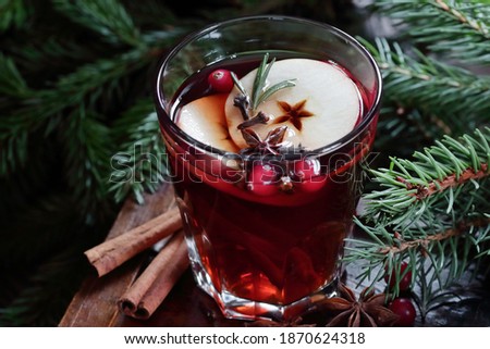 Mulled red wine with cranberry, apple and spices: anise, cinnamon, rosemary and cloves on dark moody black background and fir branches, winter christmas coctail, holiday hot drink, closeup