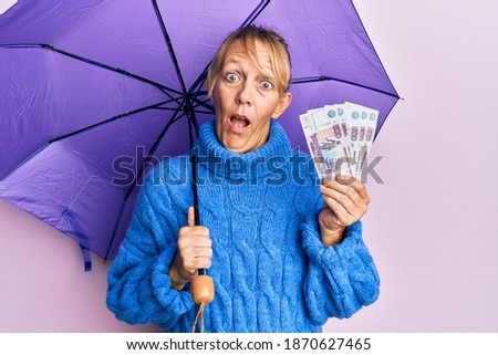 Middle age blonde woman holding umbrella and russian ruble banknotes afraid and shocked with surprise and amazed expression, fear and excited face. 