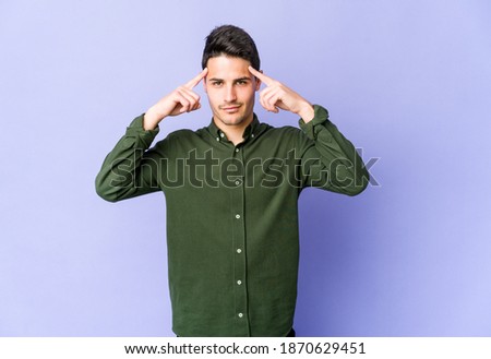 Young caucasian man isolated on purple background focused on a task, keeping forefingers pointing head.