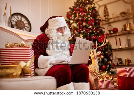 Santa Claus sitting at his home and reading email on laptop with сhristmas requesting or wish list near the fireplace and tree with gifts. New year and Merry Christmas , happy holidays concept