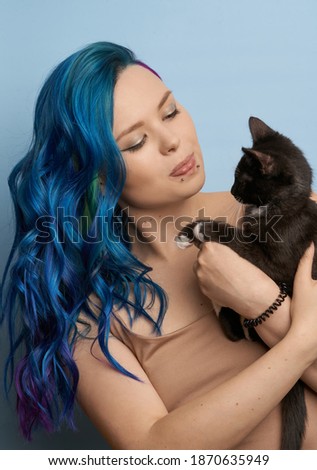 Girl with creative blue coloring and a rainbow in her hair on a blue background holds black cat. Modern minimalistic bright photography for advertising and social networks