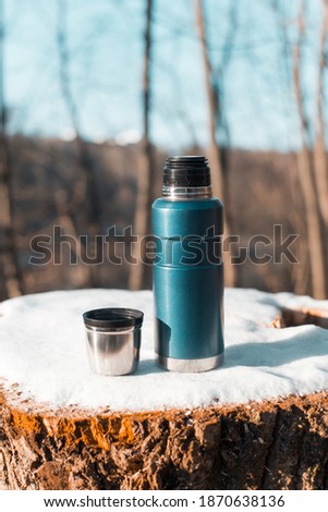 Thermos and a mug with a hot drink standing on a stump. Winter forest warming drink in a cup. Vertical photo