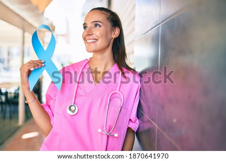 Young doctor woman holding blue cancer ribbon leaning on the wall at street of city.