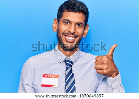 Young latin man wearing sticker with hello my name is message smiling happy and positive, thumb up doing excellent and approval sign 