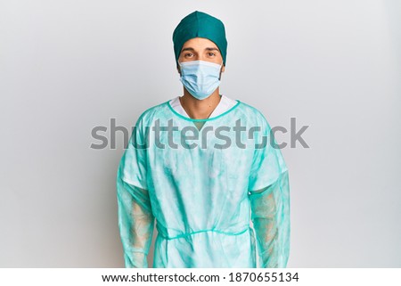 Young handsome man wearing surgeon uniform and medical mask with a happy and cool smile on face. lucky person. 
