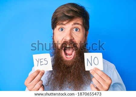 Handsome young red head man with long beard holding yes and no reminder celebrating crazy and amazed for success with open eyes screaming excited. 