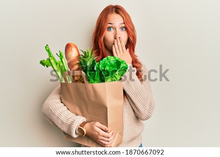 Young redhead woman holding paper bag with bread and groceries covering mouth with hand, shocked and afraid for mistake. surprised expression  Royalty-Free Stock Photo #1870667092