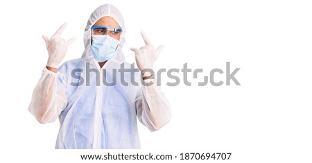 Young hispanic man wearing doctor protection coronavirus uniform and medical mask shouting with crazy expression doing rock symbol with hands up. music star. heavy concept. 