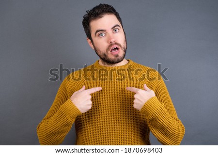Embarrassed Young handsome Caucasian man wearing yellow sweater against gray wall indicates at herself with puzzled expression, being shocked to be chosen to participate in competition.