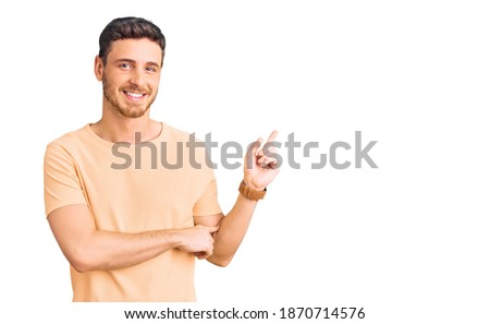 Handsome young man with bear wearing casual yellow tshirt with a big smile on face, pointing with hand and finger to the side looking at the camera. 