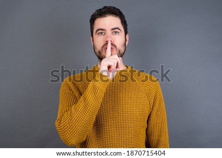 Young handsome Caucasian man wearing yellow sweater against gray wall  makes silence gesture, keeps finger over lips. Silence and secret concept.