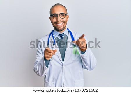 Hispanic adult man wearing doctor uniform and stethoscope pointing fingers to camera with happy and funny face. good energy and vibes. 