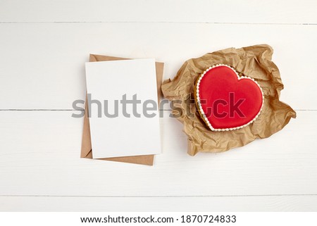 Greeting card mockup, white blank wish card with craft paper envelope and red heart shaped ginger snap cookie on wooden background. Valentine's day greeting card on white table, flat lay, top view