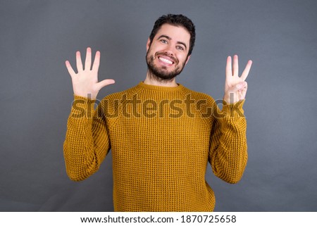 Young handsome Caucasian man wearing yellow sweater against gray wall showing and pointing up with fingers number eight while smiling confident and happy.