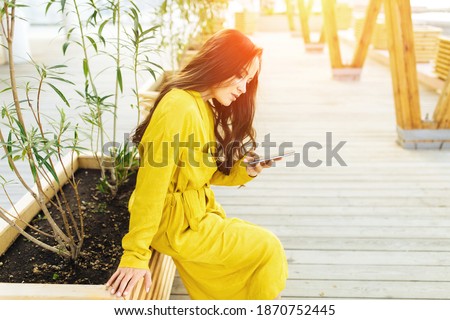 Happy girl using a smart phone in a city park sitting in yellow dress with long hair