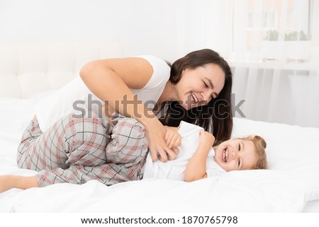 adult mother is tickling her little active daughter in bed at home, having fun, leisure activity with children.