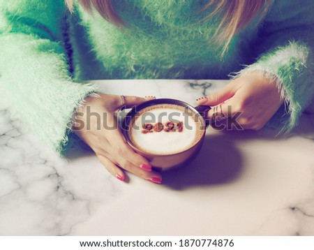 Number 2021 on frothy surface of cappuccino served in coffee cup holding by female hands with acrylic paint nail polish. Holidays food art theme for New Year 2021 celebration. (selective focus)
