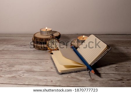 notebook or journal with a blue pencil that keeps the sheet open and an antique clock behind that tells the time by candlelight with copy space