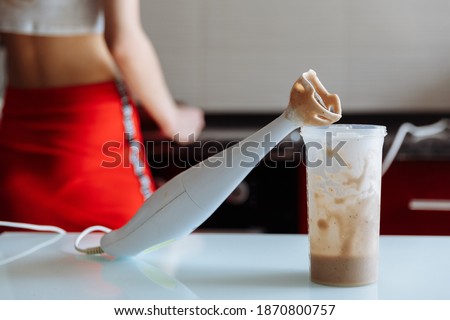 Hand blender and a shaker of Banana milkshake with the lady on background on the kitchen