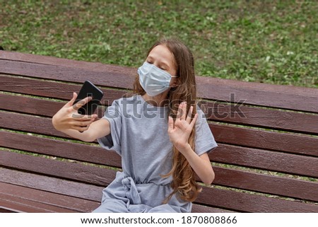 Cute girl with long blonde hair in a medical mask takes a selfie sitting on a bench in the Park.