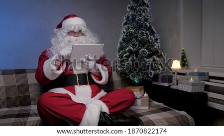 Santa Claus works remotely with a tablet online. Happy elderly man in santa claus costume smiling and looking at modern tablet sitting on sofa near christmas tree in evening in room. New Year Holidays
