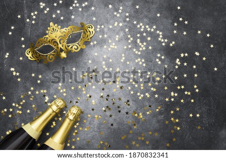 Two champagne bottles, golden carnival mask and confetti stars on grey background. Flat lay of Christmas, anniversary, New Year, Purim, Carnival celebration concept. Copy space, top view. Color 2021