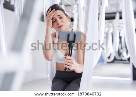 Tired unhappy brunette woman sitting at the hammock and holding smartphone at the tripod at the one hand and making video about flying yoga while holding her head with her hand and demonstrating sad