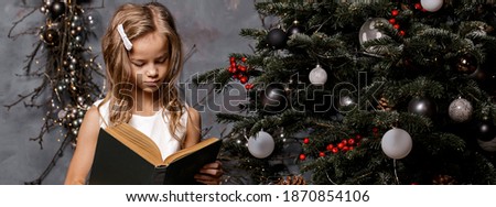 Happy child opening Christmas book. Xmas holiday concept. Girl read a book in decorated living room with Christmas tree. Banner. Copy space. Soft focus