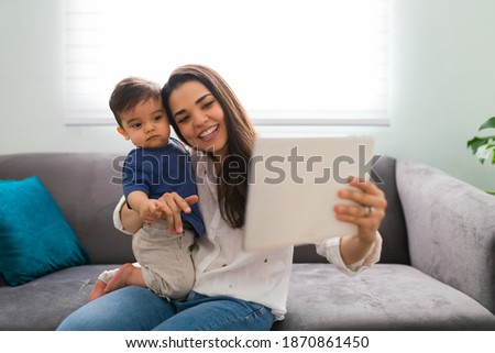 Cute toddler with her gorgeous mother using a digital tablet during a video call with family while sitting on sofa at home
