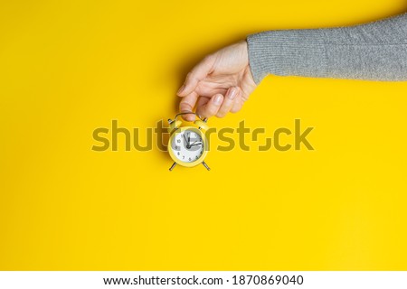 Female hand in gray clothes holding small alarm clock on yellow background. Creative banner with two colors of the year 2021 - Illuminating and Ultimate Gray. Royalty-Free Stock Photo #1870869040