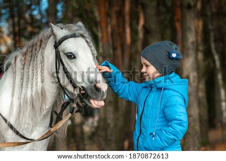 Little girl portrait, stands next to a white pony close-up on the background of nature. Jockey, epodrome, horseback riding.