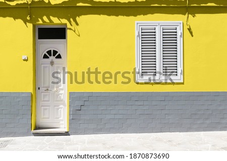 White wooden front door and white window on yellow wall with gray brick. Color trend 2021.