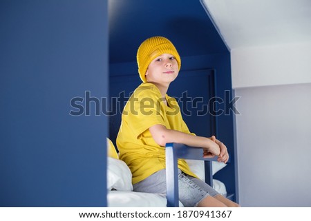Boy in yellow t-shirt and hat is spending time sitting gown on a blue two-tier bunk bed at home.