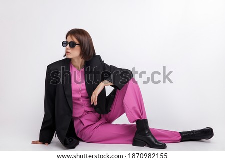 Fashion photo of a beautiful elegant young woman in a pretty oversize black jacket and pink suit,  boots, stylish sunglasses posing over white background. Bob haircut. Studio Shot